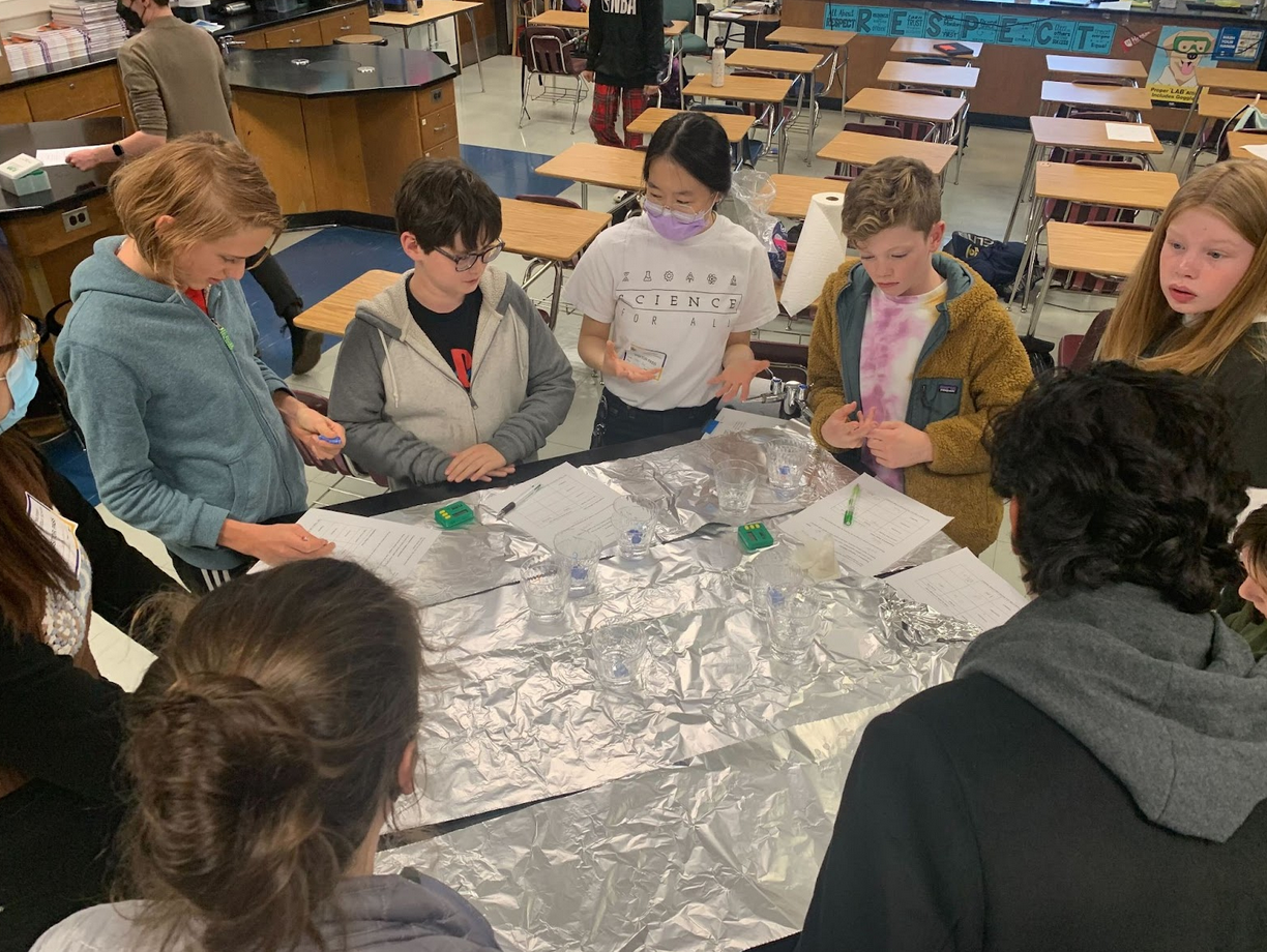 An SFA volunteer at a table covered with aluminum foil and surrounded by students discusses the day's activity. The table has clear plastic cups with a clear liquid in them, timers, and activity sheets on it. 