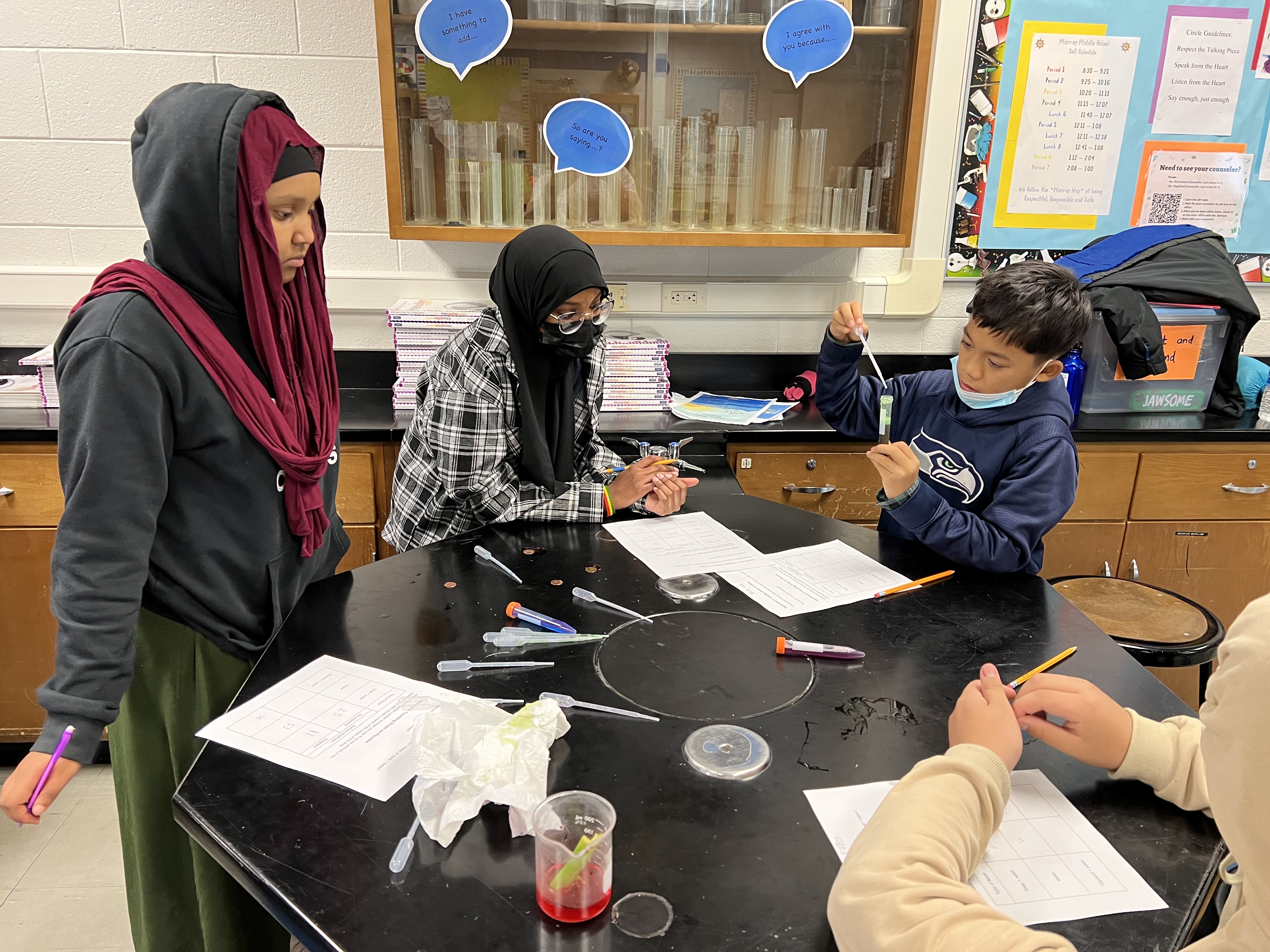 Four students surround a black table with pipettes and worksheets on it. The pipettes have different colored liquids corresponding to the liquids' surface tension. One student is holding a centrifuge tube with a green liquid (water and glycerol) in one hand and a pipette in the other.