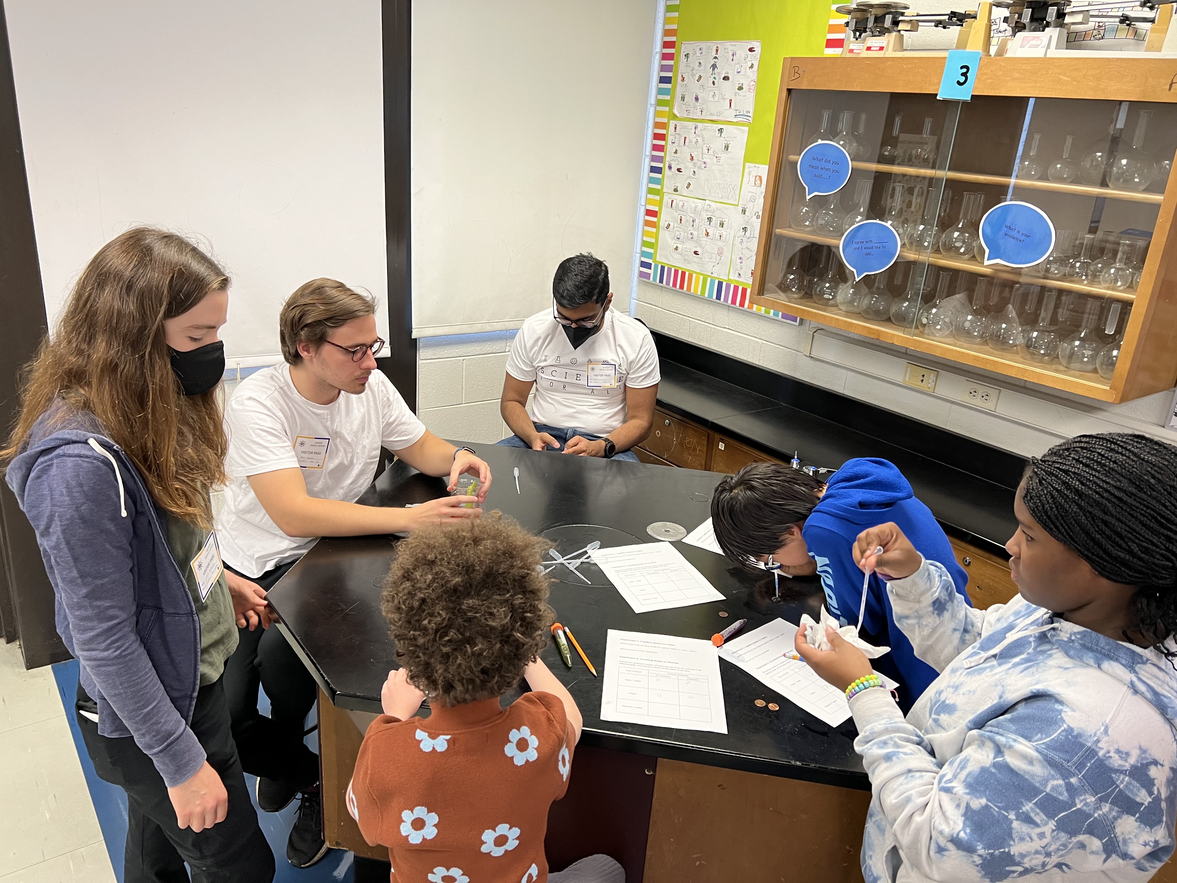 Three students and three SFA volunteers surround a black table. The students are concentrating on using pipettes to drop liquid onto pennies and count the number of drops that can fit on the penny surface. 