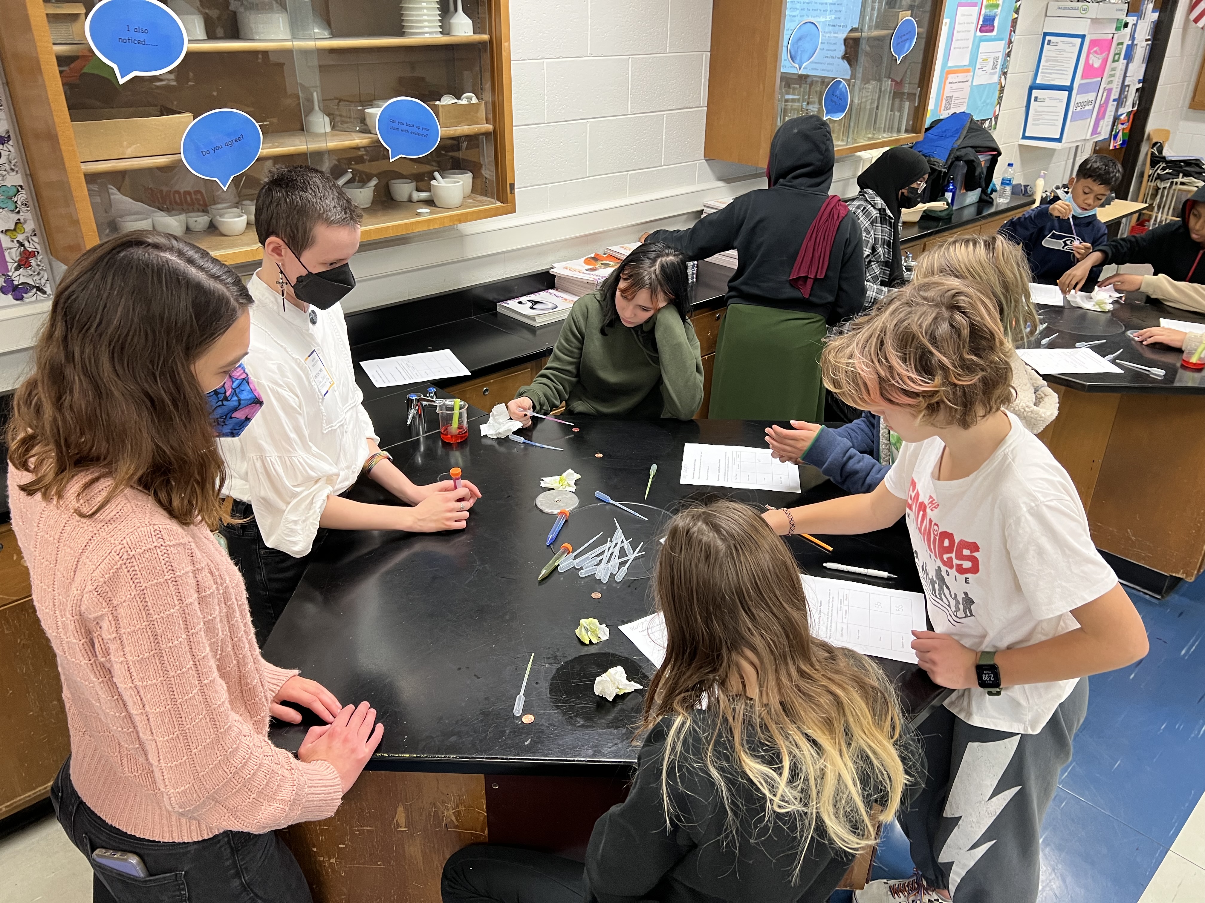 Four students and two SFA volunteers surround a black table with pipettes, centrifuge tubes with differently colored liquids, and worksheets on it. There is also a beaker with red liquid and a celery stalk in it on the table to demonstrate capillary action.  