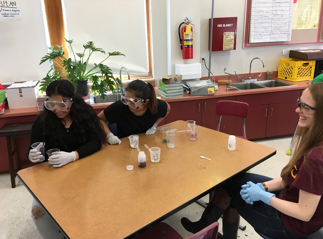 Two students with goggles and gloves on looking at mixtures in two plastic cups. An SFA volunteer sits at the opposite side of the table with glasses and gloves on. All three people in the picture are smiling.