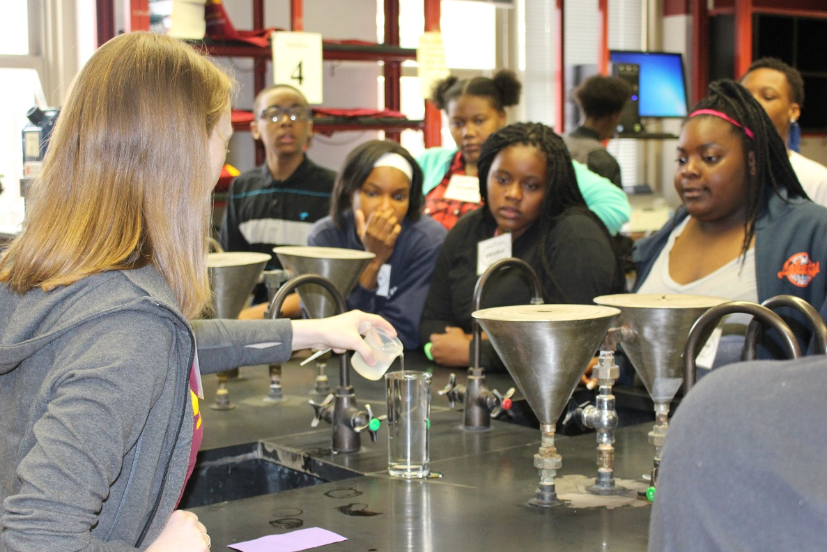 An SFA volunteer pours a clear liquid into a tall glass while a group of students watch from the other side of the lab bench. 