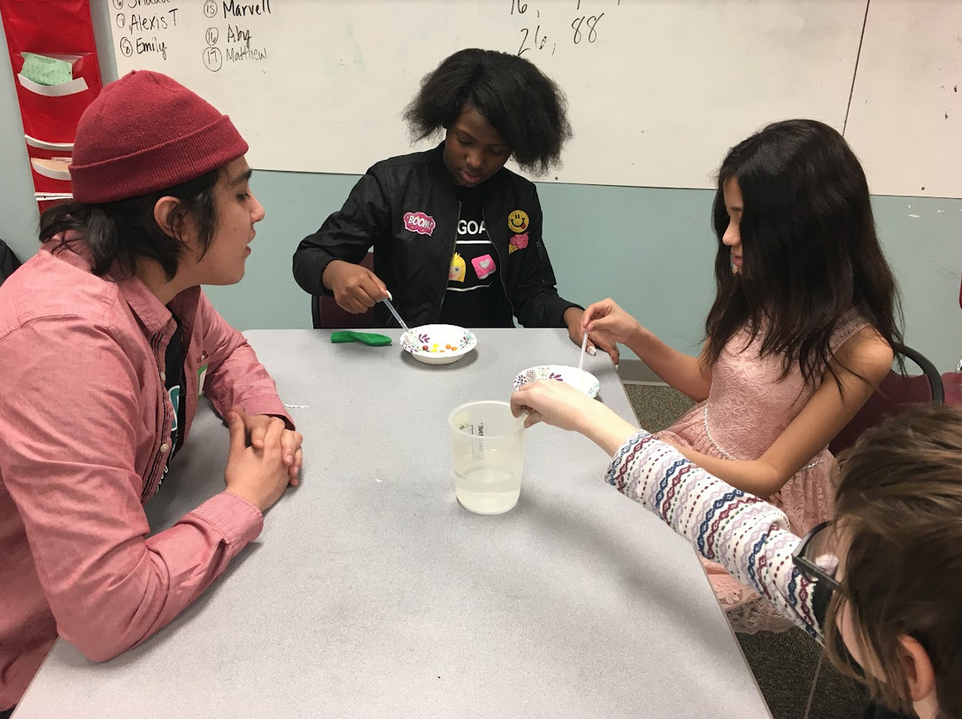 An SFA volunteer with a red shirt and a red beanie guides two students as they use pipettes over their bowls of Skittles. A third student is reaching for a beaker in front of them. 