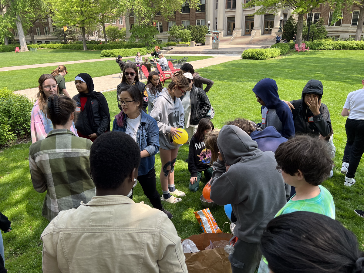 Students and SFA volunteers crowd around liquid nitrogen demonstrations on a grassy field. 