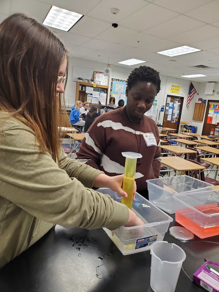 A student placing an Alka-Seltzer tablet under an inverted graduated cylinder filled with yellow liquid with the help of a volunteer. 