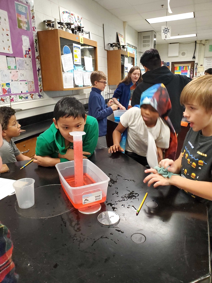 Students gathered around an inverted graduated cylinder filled with red liquid. 